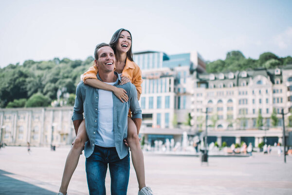 Happy young woman embracing and riding piggyback on joyful boyfriend while walking together on square during excited romantic summer tour 