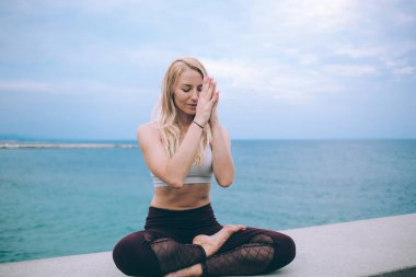 Neşeli genç bayan yoga yapıyor ve deniz kenarında eğitimden sonra kapalı gözlerle meditasyon yapıyor. Bulutlu gökyüzü elleri namaste 'de.
