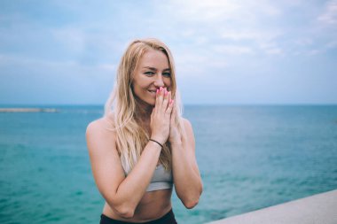 Yoga kıyafetli, neşeli, genç bir bayan. Deniz kenarında dururken avuçlarını birbirine yakın tutarak kameraya gülümsüyor.