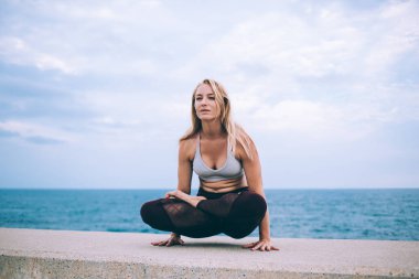 Genç, esnek, odaklanmış, siyah taytlı, beyaz saçlı, bulutlu bir günde deniz kenarında yoga yapan sarışın bir kadın.