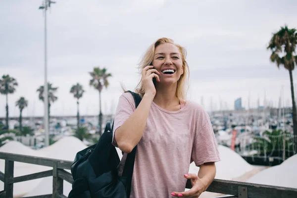 Cheerful female tourist with backpack connecting to roaming internet for calling to best friend and discuss solo travel vacations, joyful girl in casual wear enjoying international mobile talking