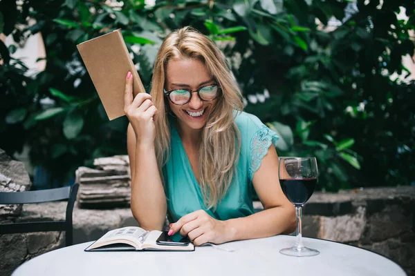 Happy successful female reader with interesting literature book taking rest in street cafe during leisure, smiling Caucasian hipster girl holding education textbook and laughing during wine drinking