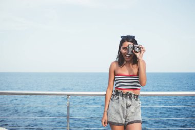 Yazlık kıyafetli kadın turist, deniz ufkunun arka planındaki setin üzerinde dikilip kameraya bakarken klasik fotoğraf makinesiyle fotoğraf çekiyor.