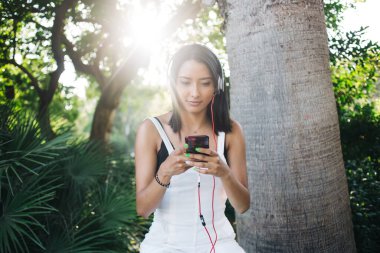 Düşünceli esmer milenyum melodisi sanatçısı akıllı telefon ve 4G bağlantısı kullanarak boş zamanlarında müzik eşliğinde eğleniyor, genç beyaz kadın açık havada cep telefonu çalma listesinden şarkı indiriyor.