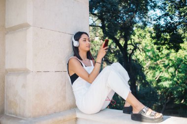 Güzel beyaz bir kadın, açık havada ses aksesuarları kullanarak online radyo dinlemek için uygulama kuruyor. Düşünceli esmer hippi kız 20 'li yaşlarda, kulaklıkta oturan cep telefonlu çalgıcı için şarkı arıyor.