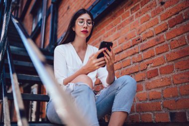 Gözlüklü, beyaz tişörtlü, ince, zeki bir bayanın altından. Kotları yırtılmış, demir merdivenlerde oturmuş şehirdeki cep telefonuyla mesajlaşıyor.