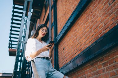 Uzun saçlı, gözlüklü, şık giyinmiş, cep telefonundan mesaj atan, yangın merdiveninde duran, kısa saçlı, Asyalı bir kadın.