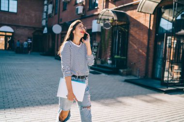 Giden neşeli, modaya uygun bir kadın elinde dizüstü bilgisayar ve defter varken cep telefonuyla arıyor.