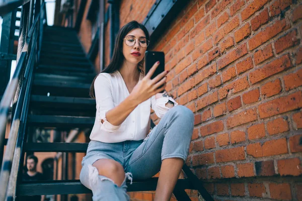 黒の建物の鉄の階段に座っているジーンズを引き裂いた白いシャツの眼鏡の焦点を当てたスマートなスリム若い女性の下から街で携帯電話で自撮り — ストック写真