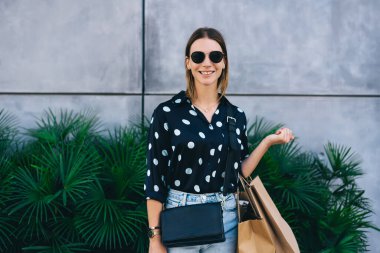 Moda kıyafetler giymiş, gözlüklü neşeli sarışının yarım boy portresi. Alışverişte mevsimlik satışların keyfini çıkarıyor. Alışveriş torbaları tutan mutlu beyaz hippi kız.