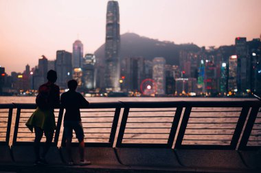 İnsanlar akşamları gökdelenlerin ışıklarına ve suya hayranlık duyarken, Hong Kong 'da dağların ardında güneş batıyor.