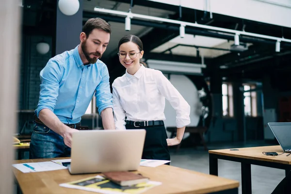 Satisfied marketing specialists in business style clothing standing near desk and working together with laptop and documents at loft creative office