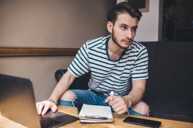 Birden fazla iş yapan adam dizüstü bilgisayarından kafasını kaldırıyor ve elinde tavayla deftere bakıyor ve odada kanepede oturuyor.