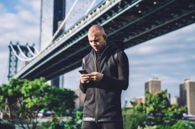 Siyah spor kıyafetleri ve kulaklıklar takan düşünceli etnik adam bulanık şehir geçmişinde güneşli bir günde telefon ekranına bakıyor.