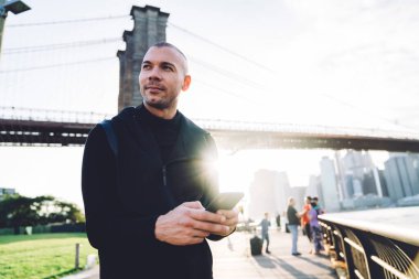 Siyah spor giyim giymiş neşeli genç bir adam metal çitlerdeki akıllı telefona dokunuyor ve şehir köprüsündeki arka planda arka plandaki ışığa bakıyor bulanık arka planda. 