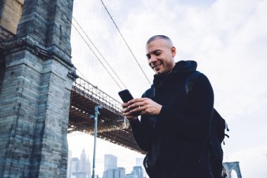 Siyah spor kıyafetli, kısa saçlı, neşeli genç bir adam. Sırt çantasıyla, kentsel arka planda, devasa bir köprünün yanındaki akıllı telefonda yazı yazıyor. 