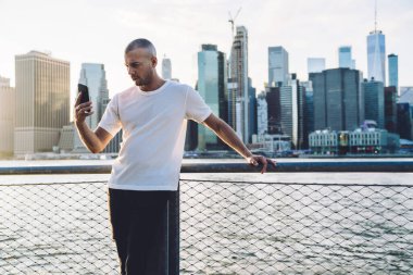 Kısa saçlı, spor elbiseli genç adam gökdelenlerin arka planında sehpada duruyor, korkuluklara tutunuyor ve akıllı telefondan selfie çekiyor.