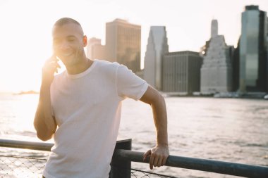 Beyaz tişörtlü genç adam güneş ışığında duruyor. Suyun yanında eskrime yaslanıyor ve telefonda gülümsüyor.