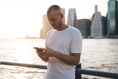 Beyaz tişörtlü genç adam gün ışığında ayakta duruyor. Suyun yanında eskrime yaslanıyor ve telefonda ciddi bir yüzle yazı yazıyor.