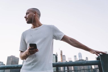 Aşağıdan gülümseyen, beyaz tişörtlü, kulaklıkla müzik dinleyen, akıllı telefon tutan metalik çitlere yaslanan ve gözlerini kaçıran genç bir adam.