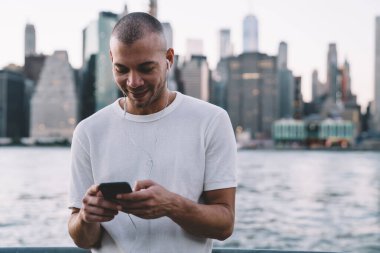 Genç gülümseyen etnik adam kulaklıkla müzik dinlerken akıllı telefon kullanıyor ve nehir ve şehrin arka planında duruyor.