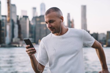 Genç gülümseyen etnik adam kulaklıkla müzik dinlerken akıllı telefon kullanıyor ve nehir ve şehrin arka planında parmaklıklara yaslanıyor.