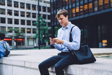 Açık mavi gömlekli ve deri çantalı genç bir adam telefonla mesajlaşıyor şehrin arka planında kablosuz kulaklıkla oturuyor.