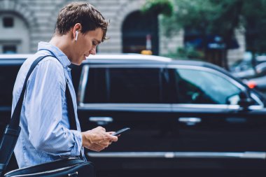 Tişörtlü, kablosuz kulaklıklı, büyük deri çantalı, sokakta arabalarla yürüyen ve gülümseyen erkek işçilerin yan görüntüsü. 