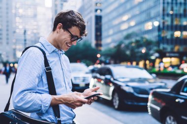 Mavi gömlekli ve gözlüklü genç erkek işçi New York caddesinde telefonla mesajlaşıyor mutlu bir şekilde gülümsüyor.