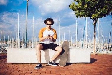 Barcelona rıhtımında çitin üzerinde oturan ve cep telefonunu karıştıran, güncel giyimli, güneş gözlüklü genç siyah saçlı bir adam.