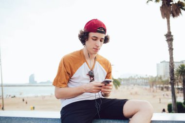 Kıvırcık saçlı genç adam, sıradan giyinmiş ve kırmızı şapkalı cep telefonuyla mesajlaşıyordu. Kulaklıkları çitin üzerinde, deniz kıyısında, Barselona 'da bulanık bir zeminde oturuyordu.