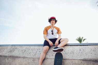 İri yapılı, modaya uygun kıyafetleri ve şapkası olan genç erkek hippi kaykaylı çitin üzerinde oturuyor ve Barselona 'daki kameraya bakıyor. 