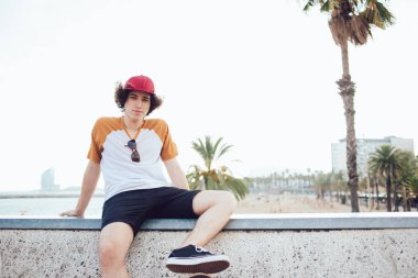 Siyah ve kıvırcık saçlı genç adam spor ayakkabı giyer ve kırmızı şapka takar kayalık çitlerde dinlenir ve Barcelona 'da arka planda tropikal deniz kıyısı olan kameraya bakar. 