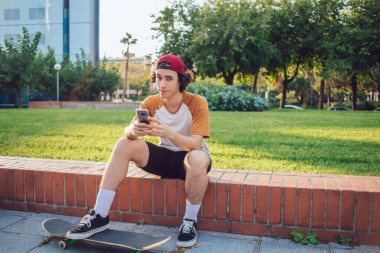 Kıvırcık saçlı erkek milenyumu şık giyiniyor spor ayakkabı ve şapka giyiyor ve kameraya bakıyor. Barselona 'da kaykayla çitte rahatlarken cep telefonunu kontrol ediyor. 