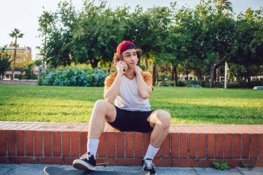 Barcelona 'da kaykayla çitin üzerinde otururken normal giyinen ve cep telefonuyla iletişim kuran kıvırcık saçlı yakışıklı bir hippi. 