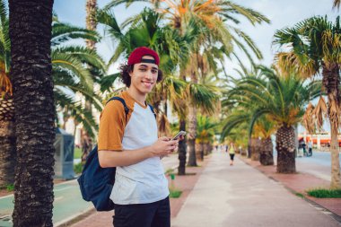 Sırt çantalı, kırmızı şapkalı, gülen ve kameraya bakan neşeli genç adam Barcelona 'da güzel bir caddede cep telefonuna bakarken. 