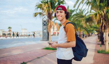 20 yaşındaki neşeli turistin yarım boy portresi güneşli Palm Caddesi 'nde duruyor ve kameraya gülümsüyor, genç milenyum kullanıcısı eğlence kaçamağı sırasında akıllı telefon cihazıyla seyahat ediyor.