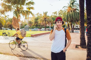 Sırt çantalı hippi bir adam telefonla konuşuyor ve sohbet ediyor. İspanya 'da palmiye ağaçları ve parlak güneş ışığı olan bir caddeye bakıyor.