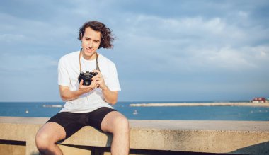 Mutlu genç erkek zamanın tadını çıkarıyor ve bulutlu gökyüzüne karşı kayalık bir çitin üzerinde oturmuş gülümsüyor ve kamerayla fotoğraf çekiyor.