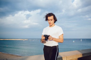 Beyaz tişörtlü genç adam bulutlu gökyüzüne karşı duruyor ve rüzgar saçlarını savururken fotoğraf makinesi kullanıyor.