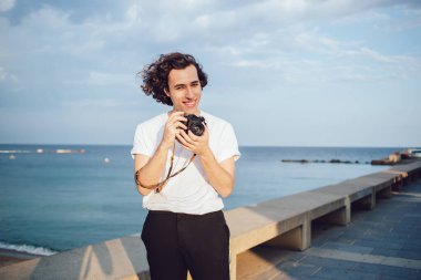 Tatmin olmuş, siyah kıvırcık saçlı, rahat giyinen, gülümseyen ve kameraya bakan genç erkek fotoğrafçı Barcelona 'da arka planda güzel bir modern rıhtıma sahip.