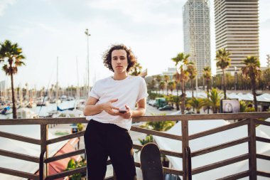 Tarz sahibi, genç, siyah saçlı, gündelik kıyafetli, ahşap çitlere yaslanmış, elinde cep telefonu olan ve Barselona 'da kameraya bakan bir adam.