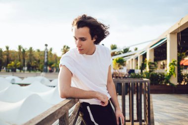 Kıvırcık saçlı, havalı kıyafetli havalı bir adamın yan görünüşü şehir kordonunda ahşap çitlere yaslanıp güneşli Barselona 'ya bakışları.