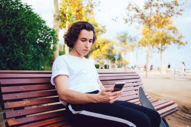 Barcelona 'da güneşli bir günde tahta bankta oturan ve cep telefonuna mesaj atan, günlük kıyafetli siyah kıvırcık saçlı adam.