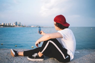 Şapkalı bir hippi mavi suyun yanında oturmuş güneşli bir günde cep telefonuyla fotoğraf çekiyor.