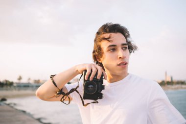 Yüze yakın fotoğraf makinesi tutan mutlu bir adam. Düşüncelere dalıp kumsalda konum alırken rüzgar kıvırcık saçlar savuruyor.