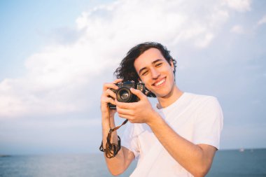 Deniz kıyısındaki hobileri sırasında kameraya gülen teknoloji ekipmanlarıyla neşeli profesyonel fotoğrafçının yarım boy portresi. Zengin 20 'li yaşlarda, medya içeriği oluşturduğu için günün tadını çıkaran bir genç.