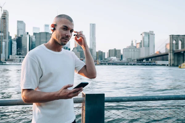 Kulaklık ve akıllı telefon tutan beyaz tişörtlü genç, kısa saçlı etnik adam çağdaş şehrin arka planında duran kameraya bakıyor.