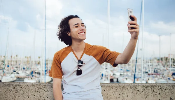 Tatmin olmuş, siyah kıvırcık saçlı, sıradan tişörtlü bir adam. Barcelona 'da arka planda parlak bir körfezle cep telefonunda selfie çekerken gülüyor.