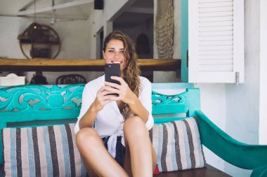 Neşeli bir kadın akıllı telefon kullanıyor rahat bir poz veriyor mavi bankta bacaklarını büküyor açık havada.